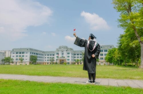 A graduate in academic regalia celebrates victory on a sunny day outdoors.