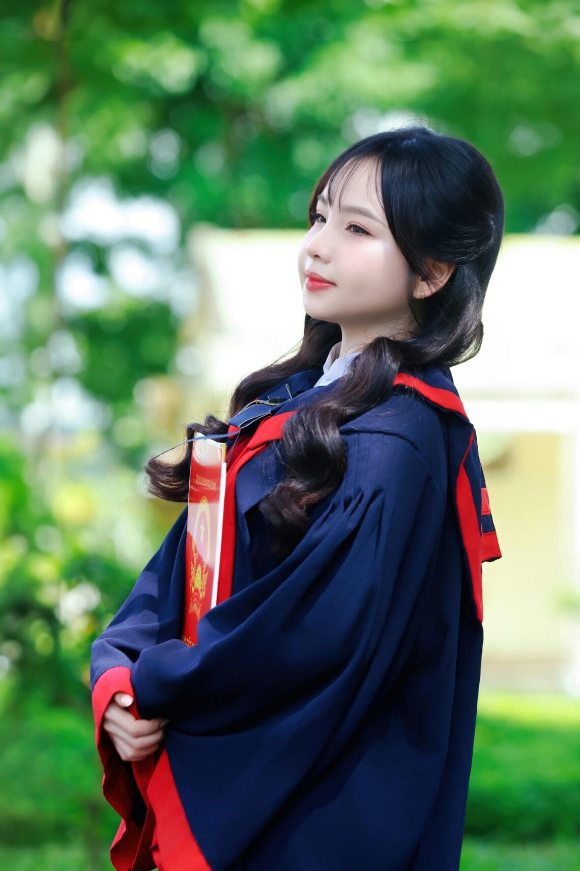 Portrait of a young graduate in a cap and gown with diploma in hand, outdoors in a lush garden.